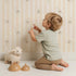 Lovely child standing in front of Whimsy Tots Milky Morning Stripes Wallpaper in Cream with milk, stripe, bottle in design.