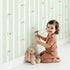 Lovely child standing in front of Whimsy Tots Milky Morning Stripes Wallpaper in Mint Green with milk, stripe, bottle in design.