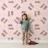 Lovely child standing in front of Whimsy Tots Moo Moo Love Wallpaper in Pink with cow, heart, bell in design.