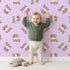 Lovely child standing in front of Whimsy Tots Moo Moo Love Wallpaper in Purple with cow, heart, bell in design.