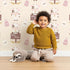 Lovely child standing in front of Whimsy Tots Moo Farm Friends Wallpaper in Cream with cow, bottle, bell, wheat, sign in design.