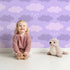 Lovely child standing in front of Whimsy Tots Dreamy Drizzle Clouds Wallpaper in Purple with cloud, star, sprinkle in design.