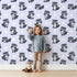 Lovely child standing in front of Whimsy Tots Playful Raccoon Parade Wallpaper in Purple with raccoon, dot in design.