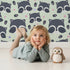 Lovely child standing in front of Whimsy Tots Winkie Raccoon Pals Wallpaper in Green with raccoon, dot, paw in design.