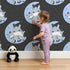 Lovely child standing in front of Whimsy Tots Moon Cuddles Zebra Wallpaper in Lavender with zebra, moon, star in design.