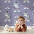 Lovely child standing in front of Whimsy Tots Rainbow Dreamy Zebra Wallpaper in Purple with zebra, rainbow, cloud, star in design.