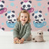 Lovely child standing in front of Whimsy Tots Cuddle Cup Panda Wallpaper in Pink with panda, cup, star, crown in design.