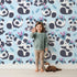 Lovely child standing in front of Whimsy Tots Tribal Hug Panda Wallpaper in Blue with panda, feather, headband, hug, leaf in design.