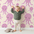Lovely child standing in front of Whimsy Tots Ocean Bubbles Octopus Wallpaper in Beige with octopus, tentacle, dot, sea in design.
