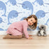 Lovely child standing in front of Whimsy Tots Bubbly Narwhal Parade Wallpaper in Pink with narwhal, horn, bubble, sea in design.