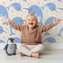 Lovely child standing in front of Whimsy Tots Bubbly Narwhal Parade Wallpaper in Beige with narwhal, horn, bubble, sea in design.