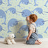 Lovely child standing in front of Whimsy Tots Bubbly Narwhal Parade Wallpaper in Mint Green with narwhal, horn, bubble, sea in design.