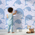 Lovely child standing in front of Whimsy Tots Bubbly Narwhal Parade Wallpaper in Lavender with narwhal, horn, bubble, sea in design.