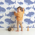 Lovely child standing in front of Whimsy Tots Whale Shark Wiggles Wallpaper in Blue with shark, whale shark, spot, sea in design.