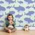 Lovely child standing in front of Whimsy Tots Whale Shark Wiggles Wallpaper in Mint Green with shark, whale shark, spot, sea in design.