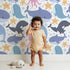 Lovely child standing in front of Whimsy Tots Ocean Buddy Parade Wallpaper in White with whale, dolphin, shark, narwhal, octopus, starfish, bubble in design.