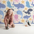 Lovely child standing in front of Whimsy Tots Ocean Buddy Parade Wallpaper in Sand with whale, dolphin, shark, narwhal, octopus, starfish, bubble in design.