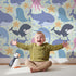 Lovely child standing in front of Whimsy Tots Ocean Buddy Parade Wallpaper in Mint Green with whale, dolphin, shark, narwhal, octopus, starfish, bubble in design.