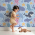 Lovely child standing in front of Whimsy Tots Ocean Buddy Parade Wallpaper in Blue with whale, dolphin, shark, narwhal, octopus, starfish, bubble in design.