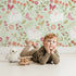 Lovely child standing in front of Whimsy Tots Meadow Hops Bunny Wallpaper in Mint Green with rabbit, butterfly, flower, leaf in design.