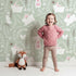 Lovely child standing in front of Whimsy Tots Bunny Hideaway Meadow Wallpaper in Mint Green with rabbit, butterfly, flower, vine in design.