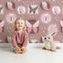Lovely child standing in front of Whimsy Tots Folk Garden Butterfly Wallpaper in Cocoa Brown with butterfly, patch, flower, dot in design.