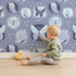 Lovely child standing in front of Whimsy Tots Folk Garden Butterfly Wallpaper in Indigo with butterfly, patch, flower, dot in design.