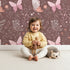 Lovely child standing in front of Whimsy Tots Wild Bloom Butterfly Wallpaper in Cocoa Rose Brown with butterfly, flower, leaf, vine in design.