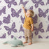 Lovely child standing in front of Whimsy Tots Celestial Wings Butterfly Wallpaper in Lilac Lavender with butterfly, moon, star, swirl in design.