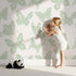 Lovely child standing in front of Whimsy Tots Celestial Wings Butterfly Wallpaper in Mint Green with butterfly, moon, star, swirl in design.