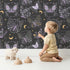 Lovely child standing in front of Whimsy Tots Starglow Garden Butterfly Wallpaper in Charcoal Gray with butterfly in design.