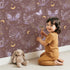 Lovely child standing in front of Whimsy Tots Starglow Garden Butterfly Wallpaper in Rosewood Pink with butterfly in design.
