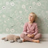 Lovely child standing in front of Whimsy Tots Floral Flutter Butterfly Wallpaper in Sage Green with butterfly, flower, leaf in design.