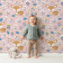 Lovely child standing in front of Whimsy Tots Meadow Cuddles Deer Wallpaper in Soft Sand with deer, flower, leaf, blossom in design.
