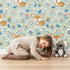 Lovely child standing in front of Whimsy Tots Meadow Cuddles Deer Wallpaper in Mint Green with deer, flower, leaf, blossom in design.