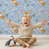 Lovely child standing in front of Whimsy Tots Meadow Cuddles Deer Wallpaper in Cornflower Blue with deer, flower, leaf, blossom in design.
