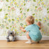 Lovely child standing in front of Whimsy Tots Bloomy Bunny Meadow Wallpaper in Buttercream with rabbit, flower, leaf, blossom in design.