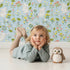Lovely child standing in front of Whimsy Tots Bloomy Bunny Meadow Wallpaper in Bluebell Sky with rabbit, flower, leaf, blossom in design.