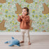 Lovely child standing in front of Whimsy Tots Blossom Bear Meadow Wallpaper in Soft Sage Green with bear, flower, leaf, blossom in design.