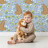 Lovely child standing in front of Whimsy Tots Blossom Bear Meadow Wallpaper in Bluebell Sky with bear, flower, leaf, blossom in design.