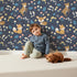 Lovely child standing in front of Whimsy Tots Meadow Mischief Fox and Deer Wallpaper in Midnight Blue with fox, deer, flower, leaf, wildflower in design.