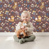 Lovely child standing in front of Whimsy Tots Meadow Mischief Fox and Deer Wallpaper in Chestnut Brown with fox, deer, flower, leaf, wildflower in design.