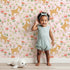 Lovely child standing in front of Whimsy Tots Spring Fawn Dance Wallpaper in Petal Pink with deer, flower, meadow, tulip in design.