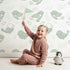 Lovely child standing in front of Whimsy Tots Tweetheart Meadow Wallpaper in Green with bird, leaf, flower, berry in design.