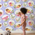 Lovely child standing in front of Whimsy Tots Garden Songbird Parade Wallpaper in Lavender with bird, branch, leaf, berry in design.