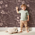 Lovely child standing in front of Whimsy Tots Woodland Silhouette Birds Wallpaper in Cocoa Brown with bird, leaf, branch, berry in design.
