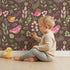 Lovely child standing in front of Whimsy Tots Bramble Songbird Garden Wallpaper in Cocoa Brown with bird, leaf, berry, flower, twig in design.