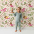 Lovely child standing in front of Whimsy Tots Blossom Branch Birds Wallpaper in Cream with bird, branch, berry, leaf in design.