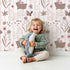 Lovely child standing in front of Whimsy Tots Meadow Hop Bunnies Wallpaper in Blush Pink with rabbit, flower, bunny, leaf, stripe in design.