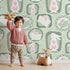 Lovely child standing in front of Whimsy Tots Framed Bunny Whimsy Wallpaper in Mint Green with rabbit, frame, bunny, flower, twig in design.